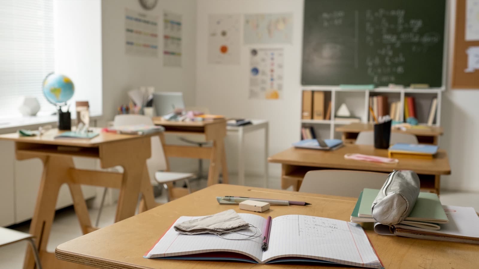 Pusta klasa: na ławce otwarty zeszyt i piórnik, w tle tablica, globus i regał z książkami