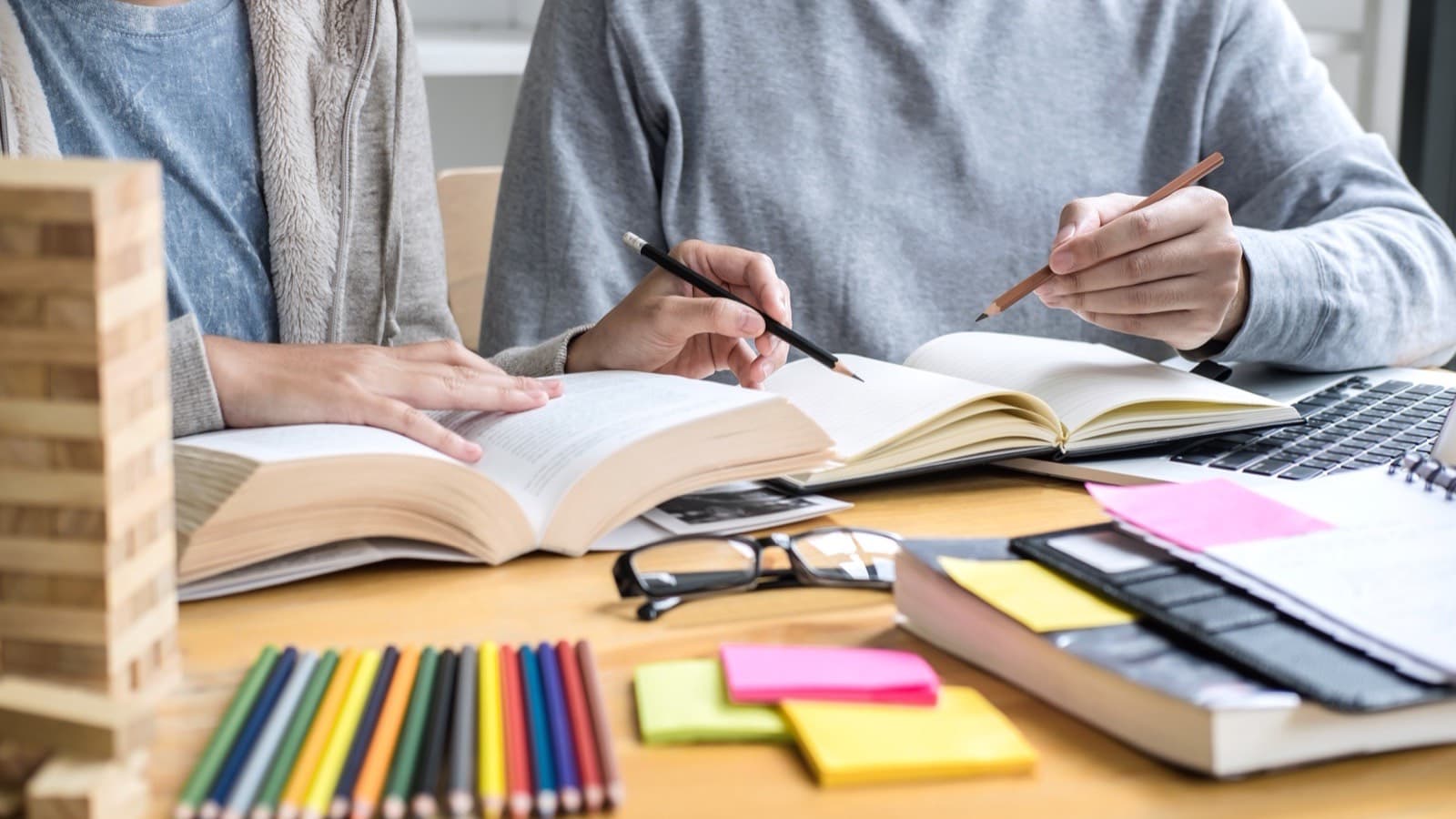 Dłonie nad otwartymi książkami, obok laptop, okulary, kredki i kolorowe karteczki na biurku